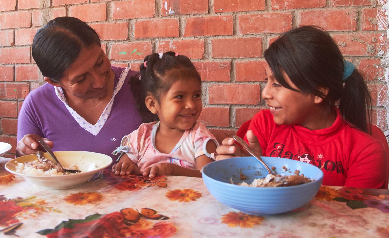 Una madre mira a sus hijas mientras comen un plato de comida. 