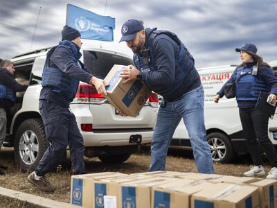 **Texto alternativo:**   El personal del Programa Mundial de Alimentos, equipado con chalecos protectores, descarga y traslada cajas selladas de asistencia alimentaria entre vehículos en una zona abierta; al fondo, bajo un cielo nublado, se ven varios coches con el logotipo del WFP y una bandera azul del WFP.