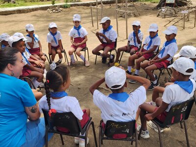 Niños con gorros blancos y pañuelos azules sentados en círculo al aire libre con personal del WFP, participando en una actividad de grupo interactiva.