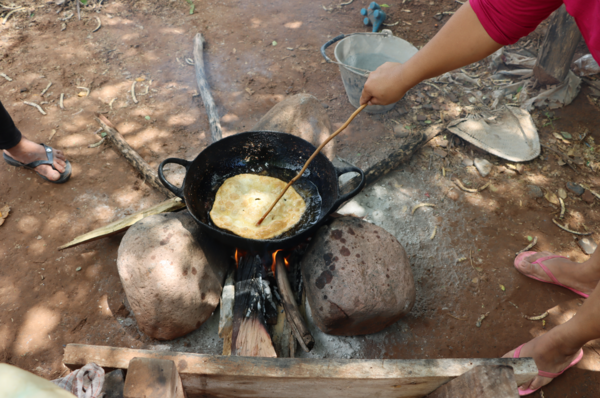 La foto muestra una sopaipilla frita en aceite en una olla sobre un fogón. La mano de una mujer usa un pequeño palo o rama para darle la vuelta y cocinarla. 