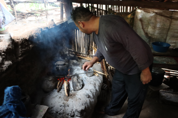 Un hombre está dentro de una cocina rural y revuelve el pescado que se está friendo en una olla sobre un fogón. Sale algo de humo proveniente de la brasa.