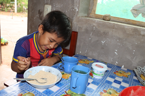 Un niño sonríe mientras está sentado a la mesa para comer su desayuno. Frente a él tiene un plato blanco lleno de maíz tostado molido y dos tazas celestes con yerba mate.