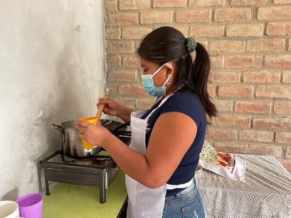 Una mujer prepara alimentos en la cocina de su casa. La mujer lleva un delantlal blanco y mascarilla o barbijo.