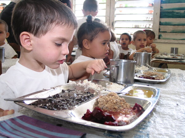 Niños y niñas en edad preescolar comen su almuerzo escolar que consiste en una bandeja metálica que contiene arroz, frijoles negros y remolacha.. 