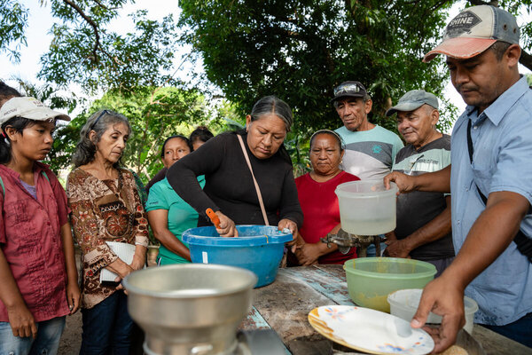 Una mujer prepara alimento para gallinas. La mujer está rodeada de otras mujeres y hombres. 