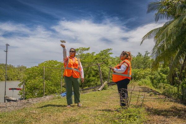 Dos mujeres con chalecos reflectores se encuentran en una zona con vegetación. Una sostiene un dron y la otra maneja el control remoto.