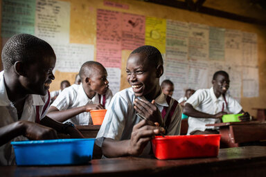 Students in school uniforms sit at wooden desks with colorful lunch containers, classroom walls covered in educational posters.
