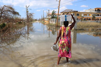 Una mujer carga en su cabeza un caja con alimentos distribuidos por WFP, mientras camina sobre una calle inundada 