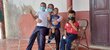 Una mujer está sentada en una silla metálica con su hijo de unos 9 años en una de sus piernas y otro hijo de unos 4 años sentado en su otra pierna. El otro hijo de unos 8 años está sentado solo en otra silla. Todos, excepto el más pequeño, llevan mascarillas o barbijos.