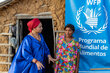 WFP Indigenous staff member in a blue cape and red headdress speaks to  woman in flowery dress outside a mud hut with WFP banner