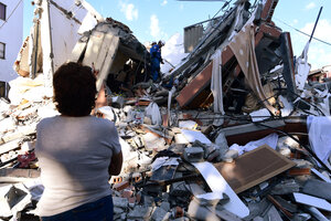 A person stands before the rubble of a collapsed building as rescue workers search through debris and twisted concrete under a blue sky.