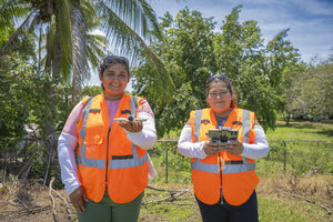 Dos mujeres con chalecos reflectores operan un dron en una zona con vegetación; una sostiene el dron y la otra, el control remoto.