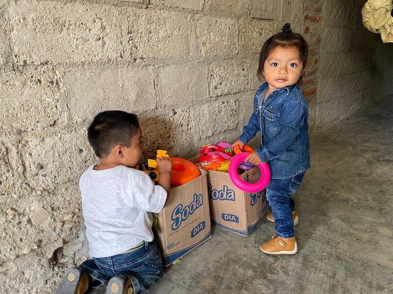Dos niños de aproximadamente 1 año de edad juegan juntos dentro de una casa. Ambos está revisando dos cajas de cartón llenas de juguetes.