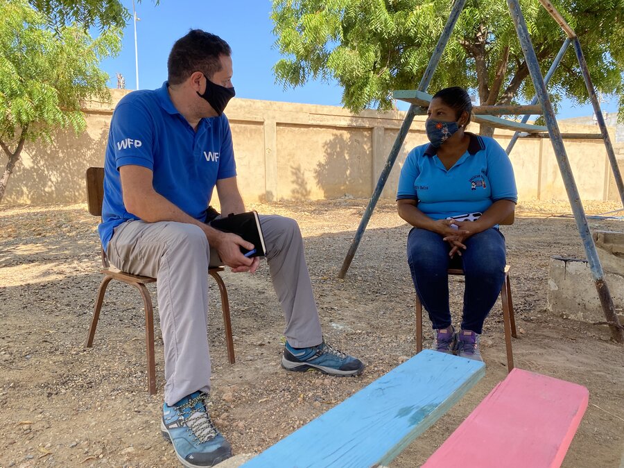 Un hombre y una mujer conversan sentados en el patio de una escuela en un dia soleado. Él es Carlos Pratts, ingeniero del Programa Mundial de Alimentos, y ella es una profesora. 