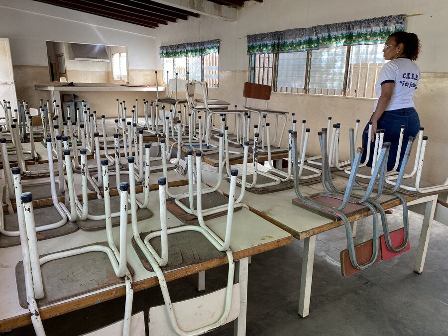 Una maestra camina dentro de un aula escolar vacía. El aula está llena pupitres y las sillas están colocadas patas arriba sobre los pupitres. 