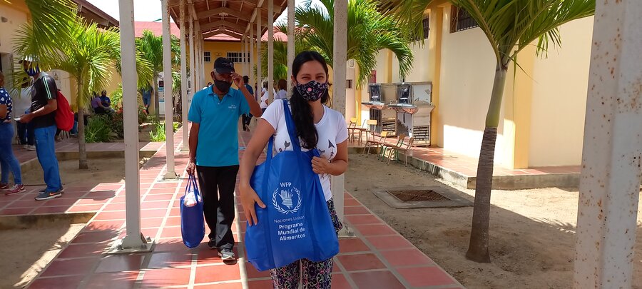 Una mujer camina por el pasillo de una escuela. Lleva colgado de su brazo derecho una bolsa azul con alimentos. La mujer lleva mascarilla o barbijo. Detrás de ella caminan otras personas. 
