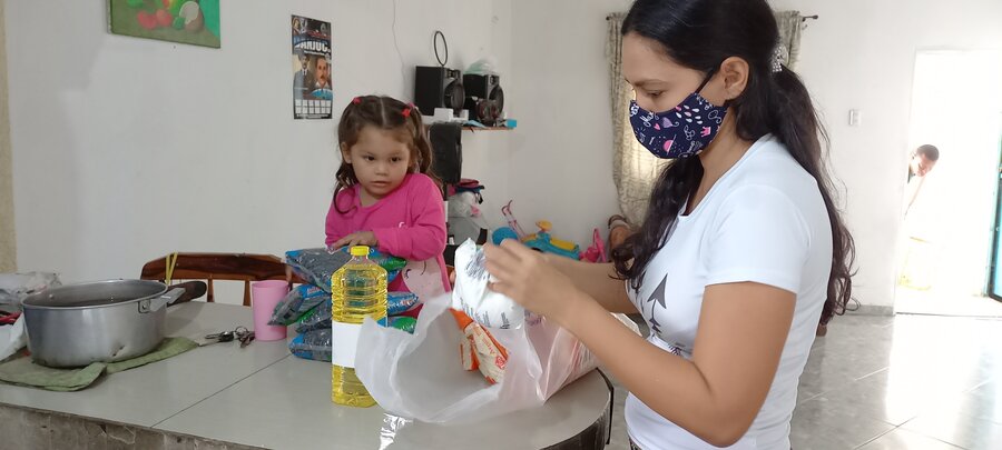 Una mujer joven sostiene en sus manos una bolsa con alimento que parece ser harina. Al fondo, sentada en la mesa una niña de unos tres años, su hija, observa. Sobre la mesa hay alimentos, como una botella de aceite, una bolsa de sal. 