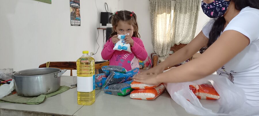 Una mujer apila unas bolsas de alimentos sobre el comedor de su casa. Al fondo, una niña de cabello largo castraño sostiene sobre su rostro una de esas bolsas.  