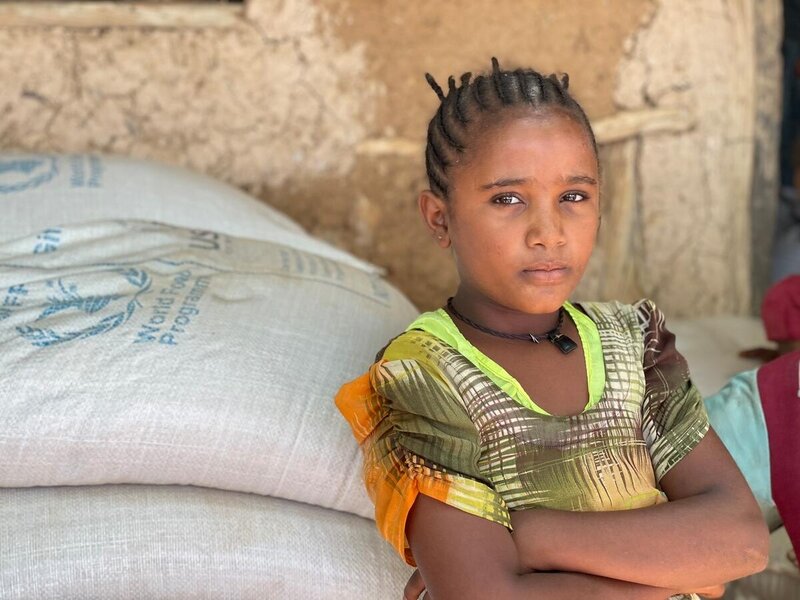 A girl stares into the camera with her arms crossed while standing next to bags of WFP food