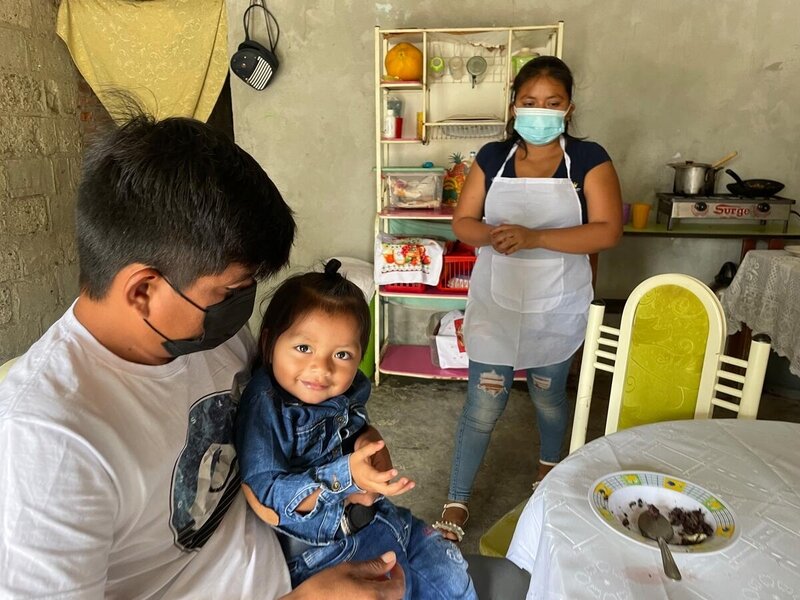 Un hombre sentado en la mesa de su casa sostiene en su regazo a su hijo mientras su esposa y madres del niño aparece al fondo con un delantal blanco.