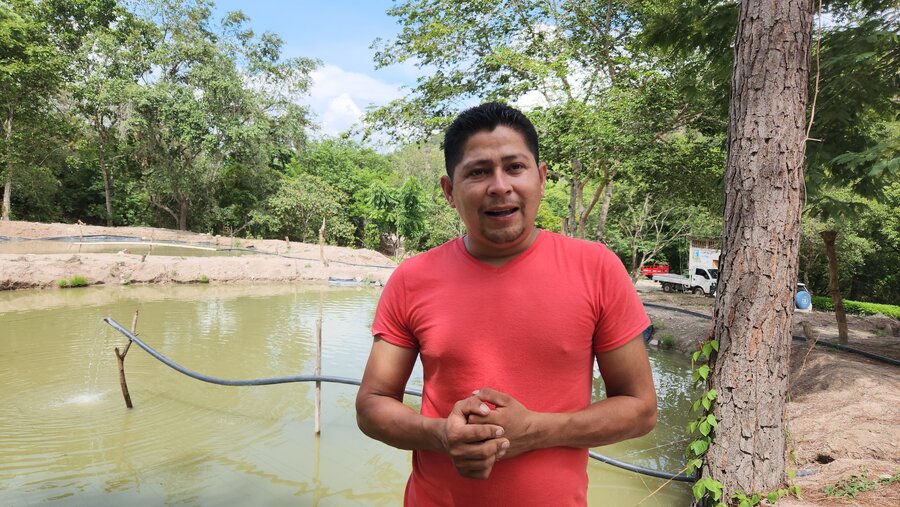 Pedro Paz habla frente a su estanque de tilapias. Pedro viste un jersey o camiseta roja.