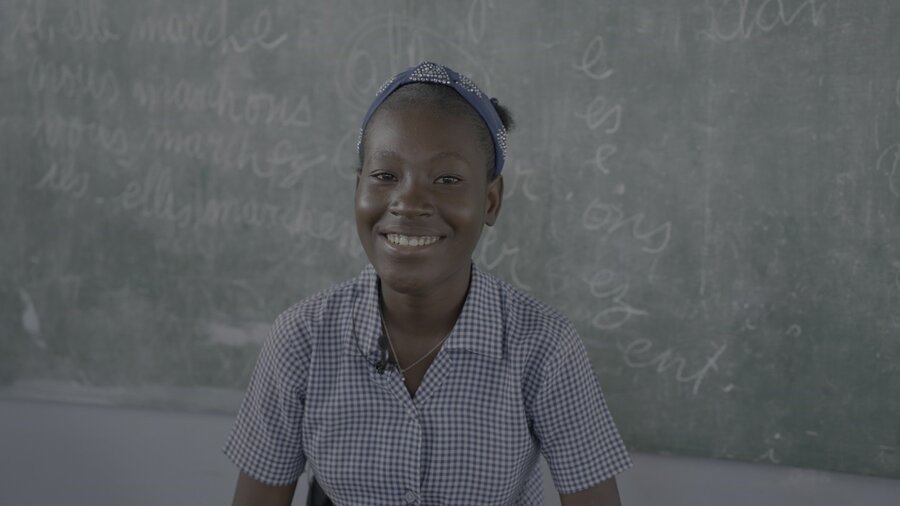 Haitian school girl in gingham shirt sits against a school blackboard