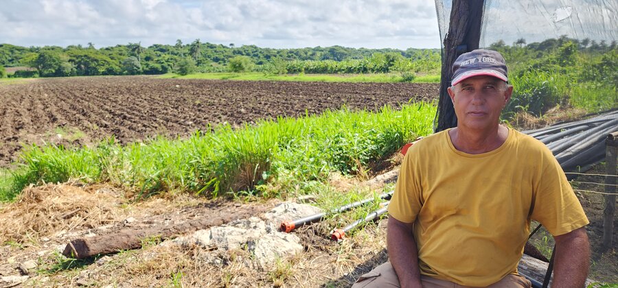 El señor Germán Broche, quien viste una camiseta amarilla y un gorra de béisbol, aparece sentado frente una casa semiprotegida y a un costado de una plantación.