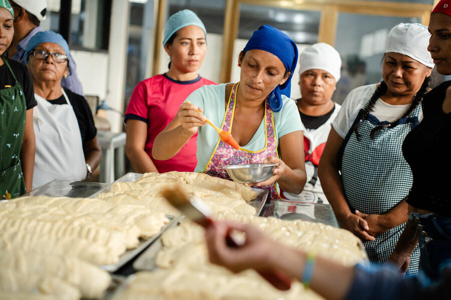 Un grupo de mujeres prepara masa de pan para hornear. Una mujer usa una brocha para poner mantequilla sobre el pan mientras las demás observan.