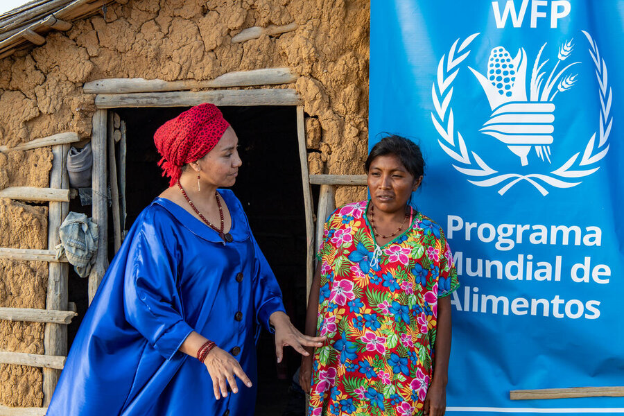 WFP Indigenous staff member in a blue cape and red headdress speaks to  woman in flowery dress outside a mud hut with WFP banner