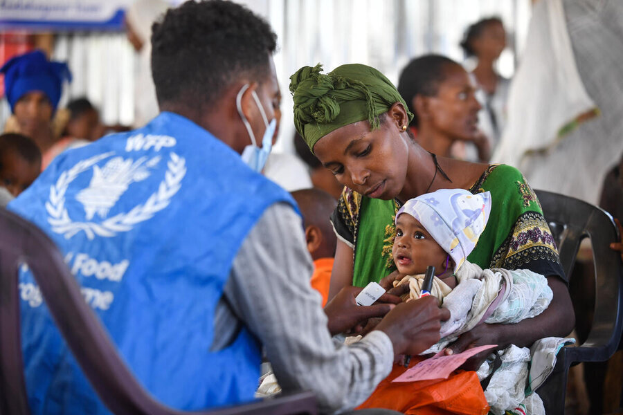 A WFP staff member in a blue vest checks a baby being held by a woman in a humanitarian aid setting, with other people visible in the background.