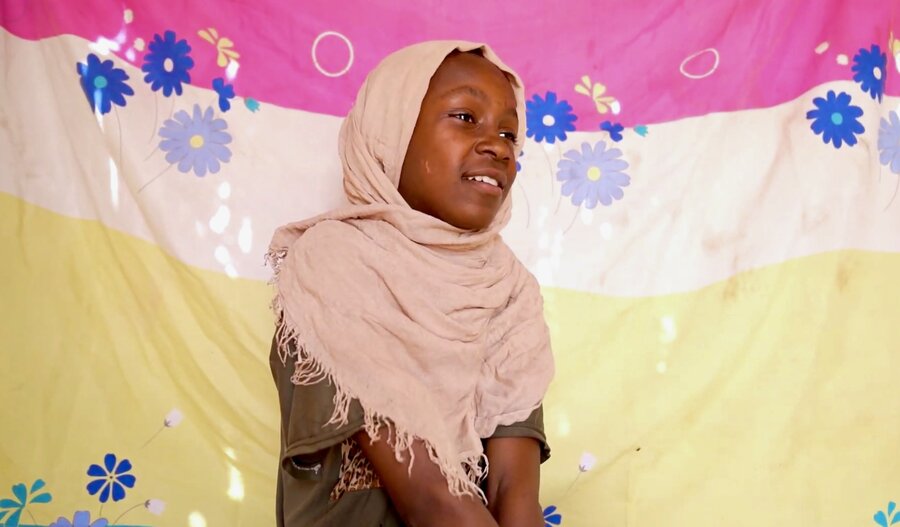 A girl in a pinkish headscarf sits in a colourful cloth tent. Photo: WFP