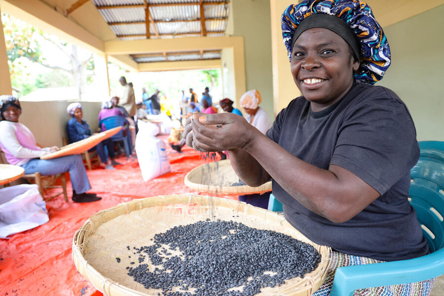 A smiling woman in a black T-shirt and colourful hat with a basket of black beans in front of her. Photo: WFP/Emily Pinna