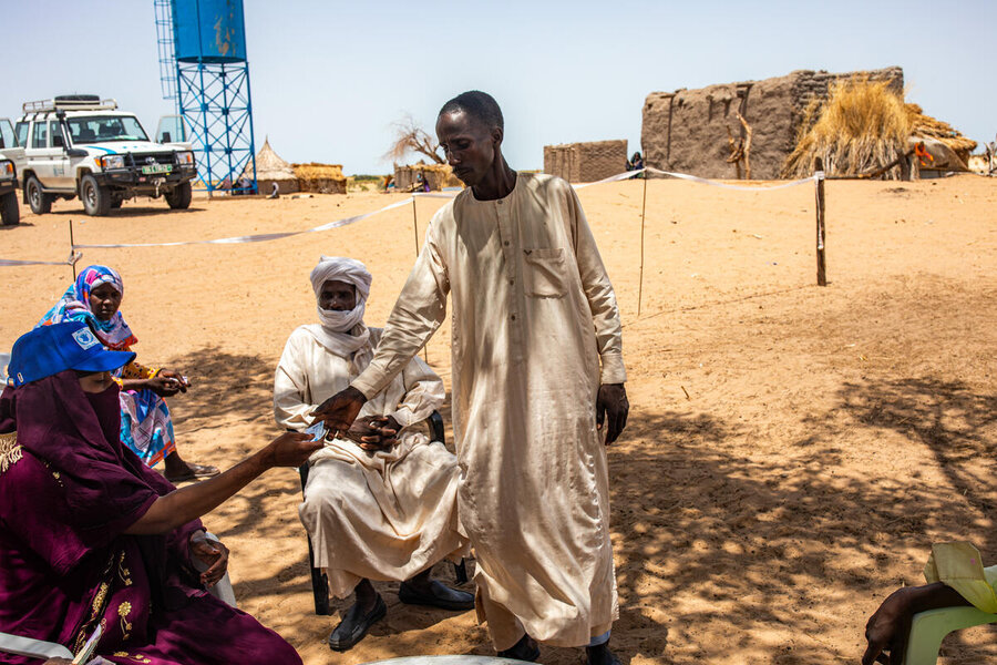 A man in a long tan robe receives WFP cash assistance from a WFP employee in Chad, while another man in a long white robe and turban watches. Photo: WFP/Aristide Dingamdoum 