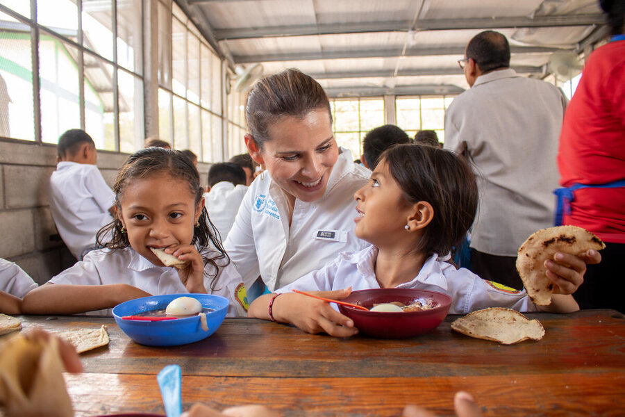A woman with a white WFP shirt chats with two little girls eating school meals in Honduras. Photo: WFP/Darlan Bautista