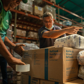Los trabajadores organizan y empaquetan productos alimenticios en un almacén, sellando y apilando cajas de aceite de girasol enriquecido para su distribución en Venezuela.