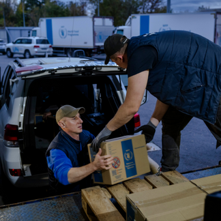 **Texto alternativo:**   Dos personas trasladan cajas de cartón selladas, con el logotipo del WFP, desde la parte trasera de un vehículo hasta una plataforma; al fondo se ven otros camiones y vehículos del WFP, lo que ilustra el apoyo logístico y la distribución de la ayuda.