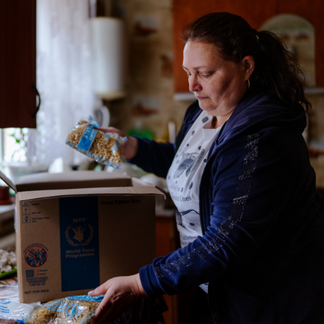 **Texto alternativo:**   Una persona en la cocina de su casa saca alimentos secos envasados de una caja de cartón con el logotipo del Programa Mundial de Alimentos y los coloca sobre la encimera, lo que muestra cómo se recibe y se organiza la asistencia alimentaria en el interior de la vivienda.