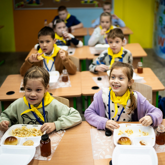 **Texto alternativo:**   Unos niños están sentados en sus pupitres en un aula de colores vivos, comiendo platos calientes servidos en bandejas con compartimentos, acompañados de pan y bebidas. Al fondo se ve a más niños sentados en sus pupitres, lo que ilustra una escena de la comida escolar.