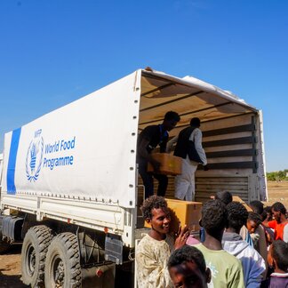 Camión del Programa Mundial de Alimentos con personal distribuyendo cajas de ayuda a las personas reunidas en el exterior; logotipo del WFP visible en el lateral del camión.