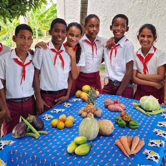 Alumnos uniformados detrás de una mesa cubierta de frutas y verduras de colores, como piña, zanahorias y berenjenas.