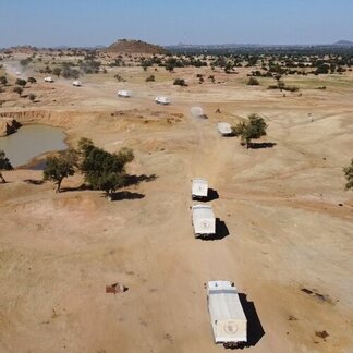 Un convoy de camiones con cubiertas blancas circula por una carretera polvorienta a través de un paisaje árido con escasos árboles y una pequeña masa de agua cerca.