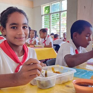 Niños uniformados comen en recipientes de plástico en una mesa amarilla dentro de un aula luminosa con grandes ventanas.