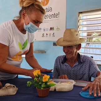 Una persona sirve comida a otra sentada en una mesa con una maceta, bajo una pancarta que promueve sistemas alimentarios sostenibles.