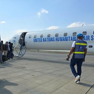 Varias personas suben a un avión del Servicio Aéreo Humanitario de la ONU en una pista, con un miembro del personal del UNHAS con chaleco caminando hacia la aeronave bajo un cielo parcialmente nublado.