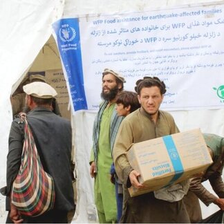 Personas reciben ayuda de WFP en una carpa con un cartel multilingüe. Una persona sostiene una caja etiquetada como Galletas Fortificadas para víctimas del terremoto.