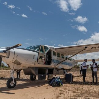 Pequeño avión en una pista de tierra con equipaje y gente de pie cerca bajo un cielo azul despejado.