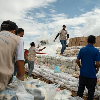Trabajadores descargan y apilan suministros de alimentos desde un barco, arrojando paquetes sobre grandes pilas de bolsas y cajas bajo un cielo nublado durante una operación de ayuda.