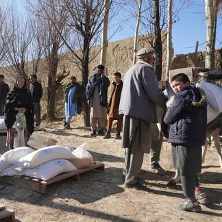 Un grupo de personas en una zona rural con árboles desnudos y un muro de barro al fondo. Varias personas están de pie, mientras que otras distribuyen sacos blancos con la etiqueta de WFP. Algunos sacos se encuentran sobre palés de madera en el suelo.